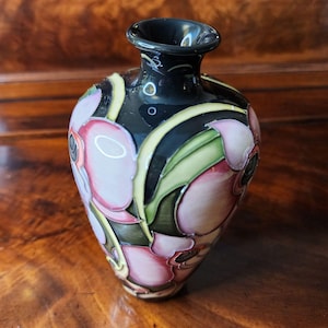 May include: A small, black vase with a flared rim, adorned with pink and white floral designs and green leaves. The vase has a glossy finish and is set against a wooden surface.