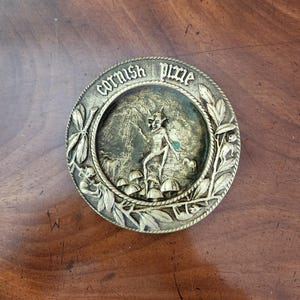 May include: A circular, antique metal plaque featuring the words "Cornish Pixie". The central image shows a pixie figure, mushrooms, and other small creatures. The plaque has an embossed design with a bronze finish, suitable for display.