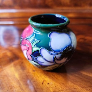 May include: A small, round ceramic vase with a dark teal rim and body. The vase is decorated with pink, white, and blue floral designs. The vase sits on a wooden surface.