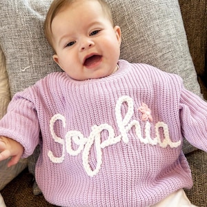 May include: A light purple knit sweater with the name "Sophia" in white script lettering. A small pink flower is next to the name. The sweater is made of a soft, textured material.