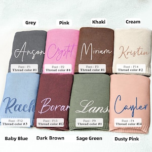 May include: A selection of personalized knit blankets in various colors, including grey, pink, khaki, and cream. Each blanket features a name embroidered in a contrasting thread color, with the font and thread color details displayed.