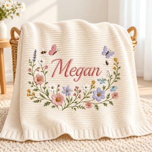 Peut inclure: Une couverture en tricot de couleur crème ornée d'une couronne de fleurs brodées dans des tons de rose, bleu et jaune. Le centre de la couronne affiche le prénom Megan en écriture cursive, agrémenté de petits papillons et abeilles brodés.