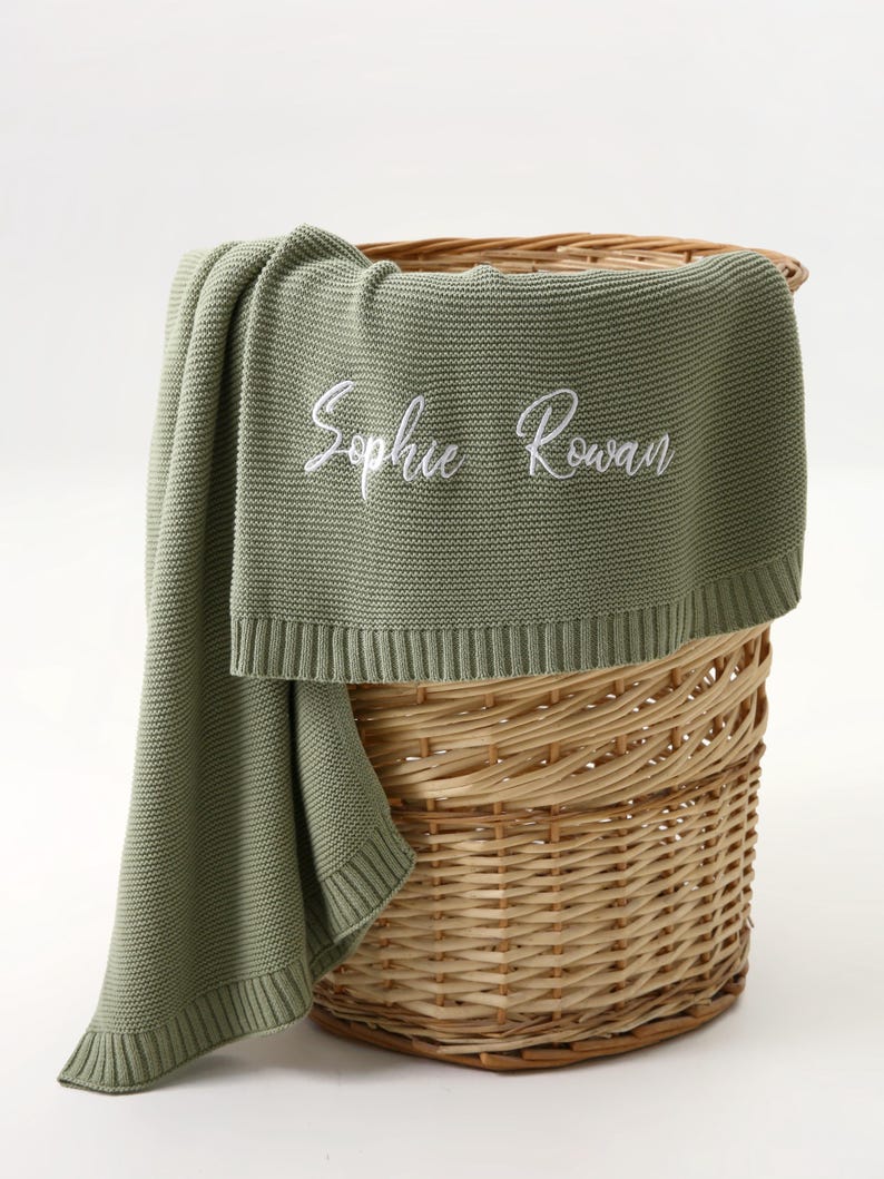 May include: A sage green knitted blanket with the embroidered name "Sophie Rowan" in white script. The blanket is draped over a woven wicker basket. The blanket has a ribbed border.