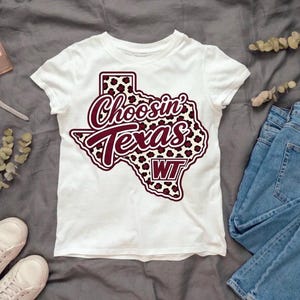 May include: A white short-sleeve t-shirt featuring a graphic of the state of Texas filled with a maroon and white leopard print pattern. The text "Choosin' Texas" is written in a maroon script font, with the letters "WT" in a maroon box below.