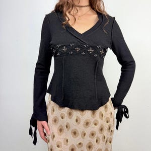 Può includere: Top nero a maniche lunghe con scollo a V e perline decorative sul petto. Il top ha una vita aderente e dettagli con fiocchi sui polsi. La modella indossa una gonna con stampa floreale.