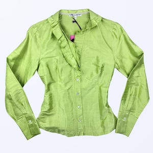 Può includere: Una camicia verde chiaro con bottoni e maniche lunghe. La camicia ha un colletto a balze e polsini con bottoni. Il tessuto ha una lucentezza sottile. Un'etichetta è attaccata alla camicia.