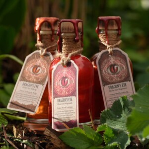 Könnte beinhalten: Drei kleine, rote Glasflaschen mit Holzstopfen und Bindfäden. Jede Flasche hat ein Etikett mit der Aufschrift "Dragon's Eye" und einer Augengrafik. Die Flaschen sind von grünem Laub und braunem Mulch umgeben.