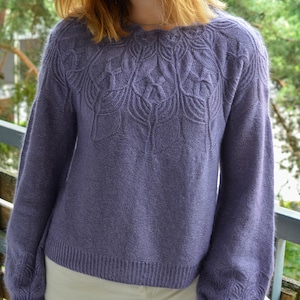 Pode incluir: Um suéter macio e roxo com um delicado padrão floral ao redor da gola. O suéter tem mangas compridas e um caimento solto.