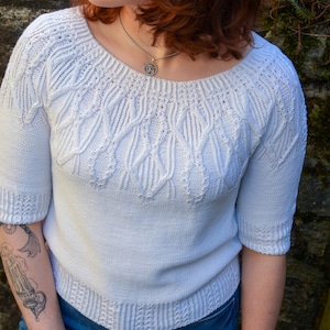 Könnte beinhalten: Ein weißer Pullover mit kurzen Ärmeln und einem strukturierten Zopfmuster auf Brust und Ärmeln. Der Pullover hat einen Rundhalsausschnitt und gerippte Bündchen und Saum. Die Person trägt eine blaue Jeans und eine silberne Halskette.