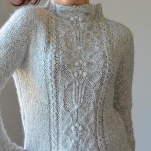 May include: A gray knitted sweater with a cable knit pattern and a high neck.