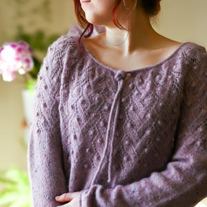 May include: A lavender-colored knitted sweater with a scoop neckline and long sleeves. The sweater features a textured pattern with small bobbles and a tie detail at the neckline. The overall design is intricate and detailed, showcasing the craftsmanship of the knit.