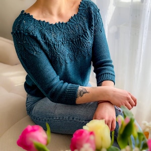 Könnte beinhalten: Ein langärmeliger Strickpullover in Blaugrün mit einem dekorativen Strukturmuster um den weiten Halsausschnitt und die Schultern, kombiniert mit einer blauen Jeans. Der Pullover hat gerippte Bündchen und ein gewelltes Detail am Kragen.