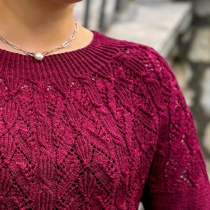 Könnte beinhalten: Nahaufnahme eines weinroten Strickpullovers mit Rundhalsausschnitt und aufwendigen blattartigen Mustern. Der Pullover hat ein strukturiertes Design und besteht aus einem weichen Material. Eine silberne Halskette mit Perlenanhänger ist sichtbar.