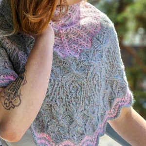 Puede incluir: Un chal de punto gris claro y rosa con un patrón de encaje. El chal está drapeado sobre los hombros de una persona y se lleva con una camisa blanca y unos vaqueros azules.