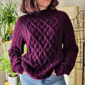 May include: A deep purple cable knit sweater with a diamond pattern. The sweater has long sleeves and a crew neck.