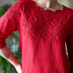 Könnte beinhalten: Ein leuchtend roter Strickpullover mit einem wellenförmigen Ausschnitt und aufwendigen Mustern an Passe und Saum. Das langärmlige Kleidungsstück hat ein strukturiertes Design, das die Handwerkskunst des Strickens zeigt.