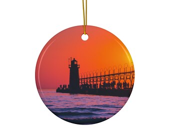 May include: A round ceramic bauble depicting a silhouette of a lighthouse and pier at sunset. The sky is a vibrant orange and yellow, with the water a deep purple.