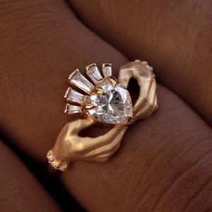 May include: A gold-colored ring featuring a heart-shaped diamond center stone, surrounded by smaller baguette-cut diamonds. The ring's design includes two hands holding the central stone. The word "Jewelasy" is at the bottom.