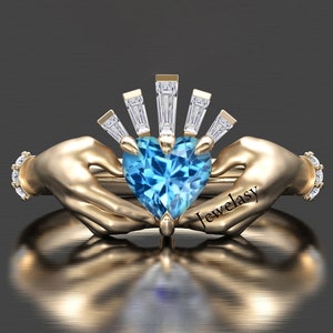 May include: A gold-colored ring featuring a heart-shaped, light blue gemstone held by two hands. The ring has a sunburst design above the heart and small diamonds on the band. The word "Jewelasy" is printed on the ring.
