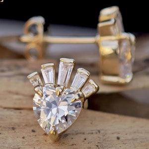 May include: Gold-toned stud earrings with a heart-shaped, clear gemstone center, surrounded by smaller, rectangular gemstones. The earrings are set on a wooden surface with a dark background. The brand name "Jewelasy" is visible in the upper left corner.