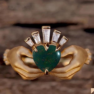 Op de afbeelding: Een gouden ring met twee handen die een groene hartvormige steen vasthouden. De steen is omgeven door een halo van heldere baguette-vormige stenen.