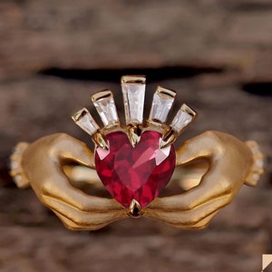 May include: A gold ring featuring two hands clasping a red heart-shaped gemstone. The ring is decorated with a crown of small clear gemstones.
