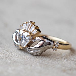 Może przedstawiać: Dwukolorowy pierścionek Claddagh z diamentem w kształcie serca. Pierścionek przedstawia dłonie trzymające serce, zwieńczone koroną. Oprawa jest złota, ze srebrną oprawą serca i dłoni.