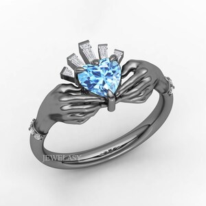 Può includere: Un anello di Claddagh con una gemma a forma di cuore, azzurra, tenuta da due mani. L'anello ha una fascia grigio scuro ed è adornato con piccole gemme trasparenti. La parte superiore dell'anello presenta un design a raggiera.