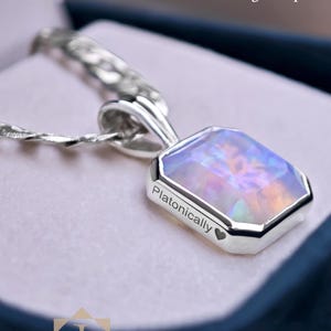 Op de afbeelding: Een zilveren hanger met een iriserende, rechthoekige edelsteen. De hanger is gegraveerd met het woord "Platonically" en een hartsymbool. De ketting wordt gepresenteerd in een donkerblauwe doos. Tekst op de afbeelding zegt "Deze hanger is volledig aanpasbaar."