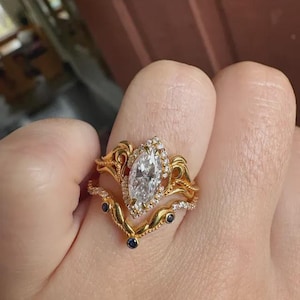 May include: A gold engagement ring set with a marquise-cut diamond centre stone, encircled by smaller diamonds. The set includes a curved band with small blue gemstones and a matching band with delicate detailing.
