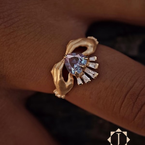 Könnte beinhalten: Ein goldfarbener Ring mit einem birnenförmigen, hellblauen Edelstein, akzentuiert mit Baguette-Schliff-Steinen. Das Design des Rings umfasst stilisierte Hände, die den zentralen Stein halten. Der Markenname "Jewelasy" ist sichtbar.