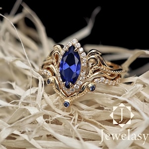 May include: A gold ring set with a large, marquise-cut, deep blue gemstone and smaller white gemstones. The ring has an ornate, swirling design with small blue gemstones. The word "Jewelasy" is in the bottom right corner.
