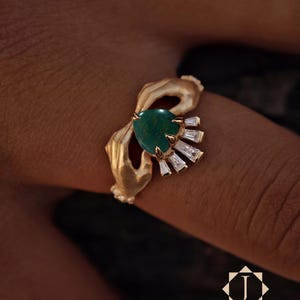 以下が含まれることがあります： ハート型の濃い緑色の宝石とバゲットカットのクリアストーンがアクセントになったゴールドトーンのリング。リングのデザインには、様式化された手のモチーフが含まれています。「Jewelasy」のロゴが右下に表示されています。