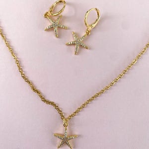 Puede incluir: Un conjunto de collar y pendientes de oro con un colgante de estrella de mar y pendientes a juego. La estrella de mar est&aacute; adornada con piedras transparentes brillantes.