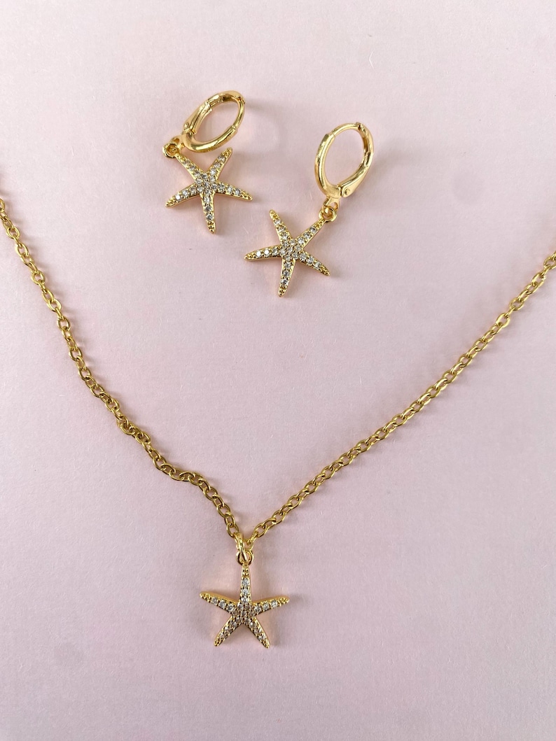 Puede incluir: Un conjunto de collar y pendientes de oro con un colgante de estrella de mar y pendientes a juego. La estrella de mar est&aacute; adornada con piedras transparentes brillantes.