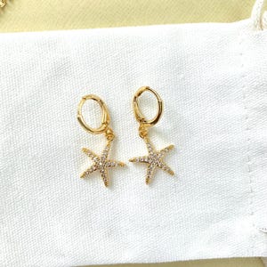 Puede incluir: Un par de pendientes de aro dorados con un peque&ntilde;o encanto de estrella de mar brillante colgando de cada aro.