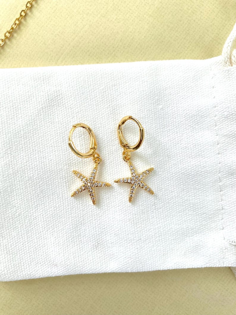 Puede incluir: Un par de pendientes de aro dorados con un peque&ntilde;o encanto de estrella de mar brillante colgando de cada aro.