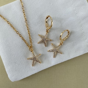 Puede incluir: Un conjunto de collar y pendientes de oro con un colgante de estrella de mar brillante y pendientes a juego. Las joyas se muestran sobre un fondo de tela blanca.