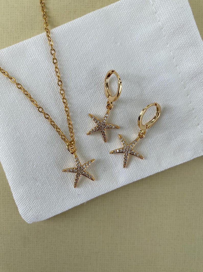 Puede incluir: Un conjunto de collar y pendientes de oro con un colgante de estrella de mar brillante y pendientes a juego. Las joyas se muestran sobre un fondo de tela blanca.