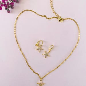 Puede incluir: Un conjunto de collar y pendientes de oro con un colgante de estrella de mar brillante y pendientes a juego. Las joyas est&aacute;n sobre un fondo rosa.