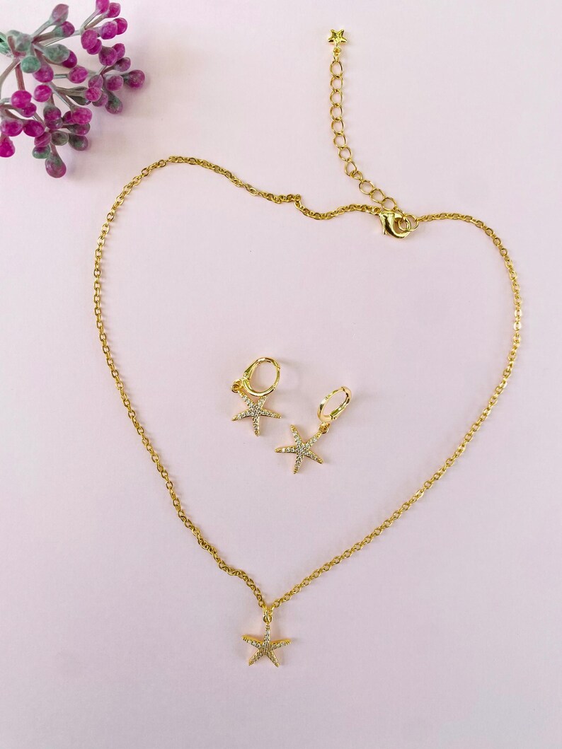 Puede incluir: Un conjunto de collar y pendientes de oro con un colgante de estrella de mar brillante y pendientes a juego. Las joyas est&aacute;n sobre un fondo rosa.