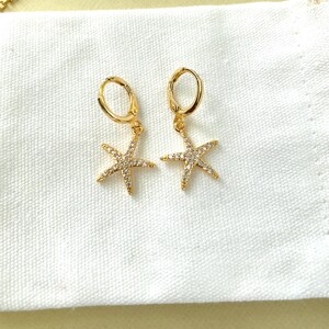 Puede incluir: Pendientes de aro dorados con un colgante de estrella de mar. La estrella de mar est&aacute; incrustada con brillantes transparentes.