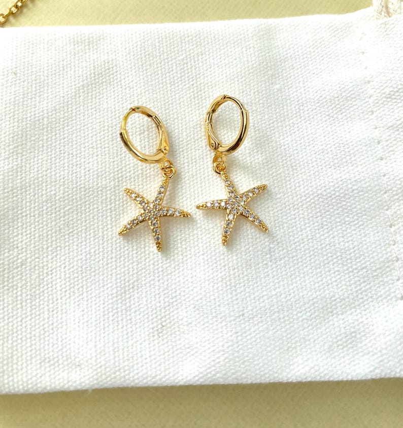 Puede incluir: Pendientes de aro dorados con un colgante de estrella de mar. La estrella de mar est&aacute; incrustada con brillantes transparentes.
