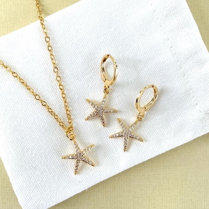 May include: A gold necklace and earrings set featuring a sparkling starfish pendant and matching earrings. The jewellery is displayed on a white fabric background.