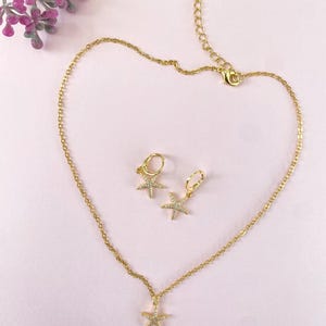 Puede incluir: Un conjunto de collar y pendientes de oro con un colgante de estrella de mar brillante y pendientes a juego. Las joyas est&aacute;n sobre un fondo rosa.