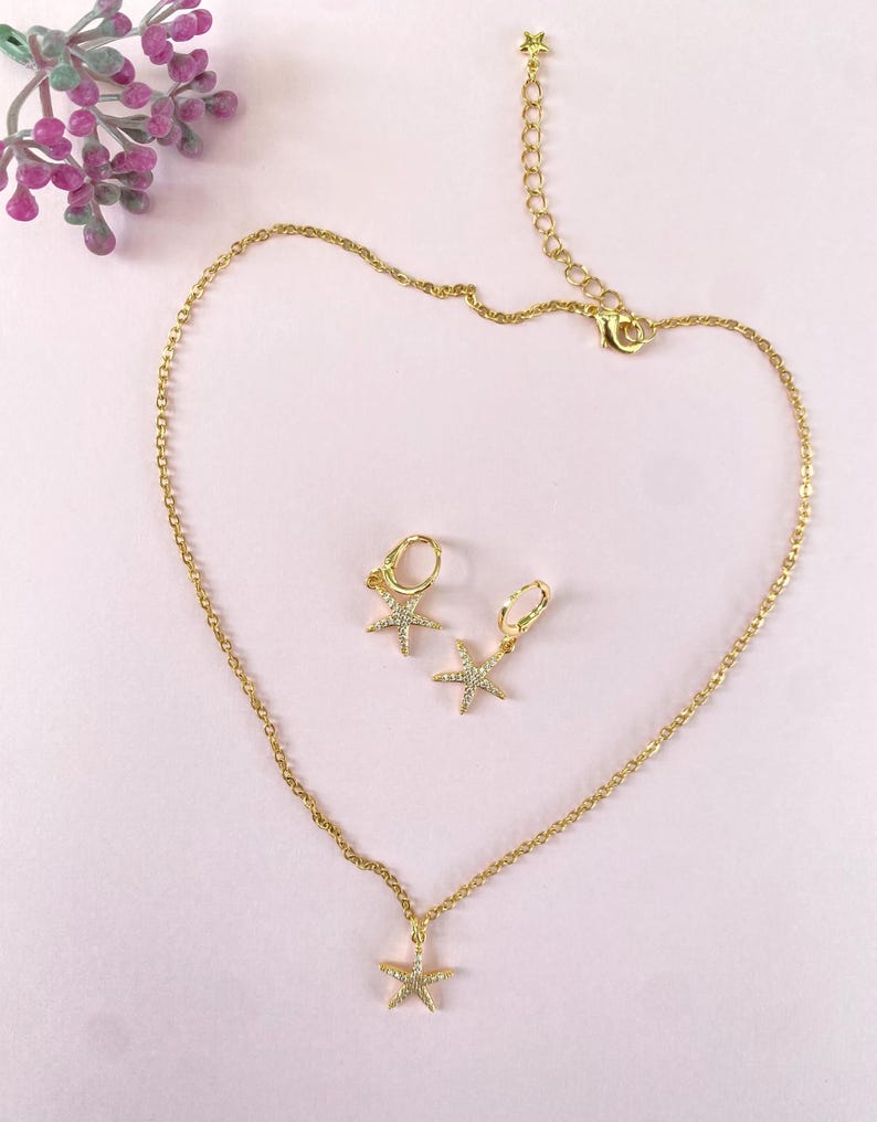 Puede incluir: Un conjunto de collar y pendientes de oro con un colgante de estrella de mar brillante y pendientes a juego. Las joyas est&aacute;n sobre un fondo rosa.