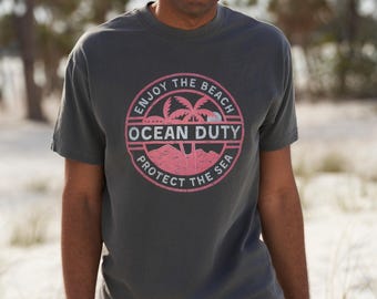 May include: A dark gray t-shirt with a pink and white graphic print of a palm tree and the text "Enjoy the Beach Ocean Duty Protect the Sea".