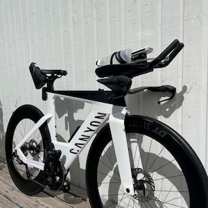 May include: A white and black Canyon triathlon bicycle featuring deep-section DT Swiss carbon wheels, disc brakes, and an aerodynamic frame design. The bike is equipped with aero handlebars, a water bottle mounted between the bars, and a saddle bag.