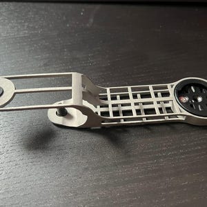 May include: A light gray bicycle computer mount with a black computer holder. The mount has a rectangular grid design and a circular end with a black computer mount. The mount is made of metal and has a bolt on each end.