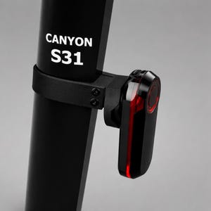 May include: Close-up of a black bicycle component with the text "CANYON S31" in white. A black strap and bracket secure a red and black light to the component. The light has a red circular design.